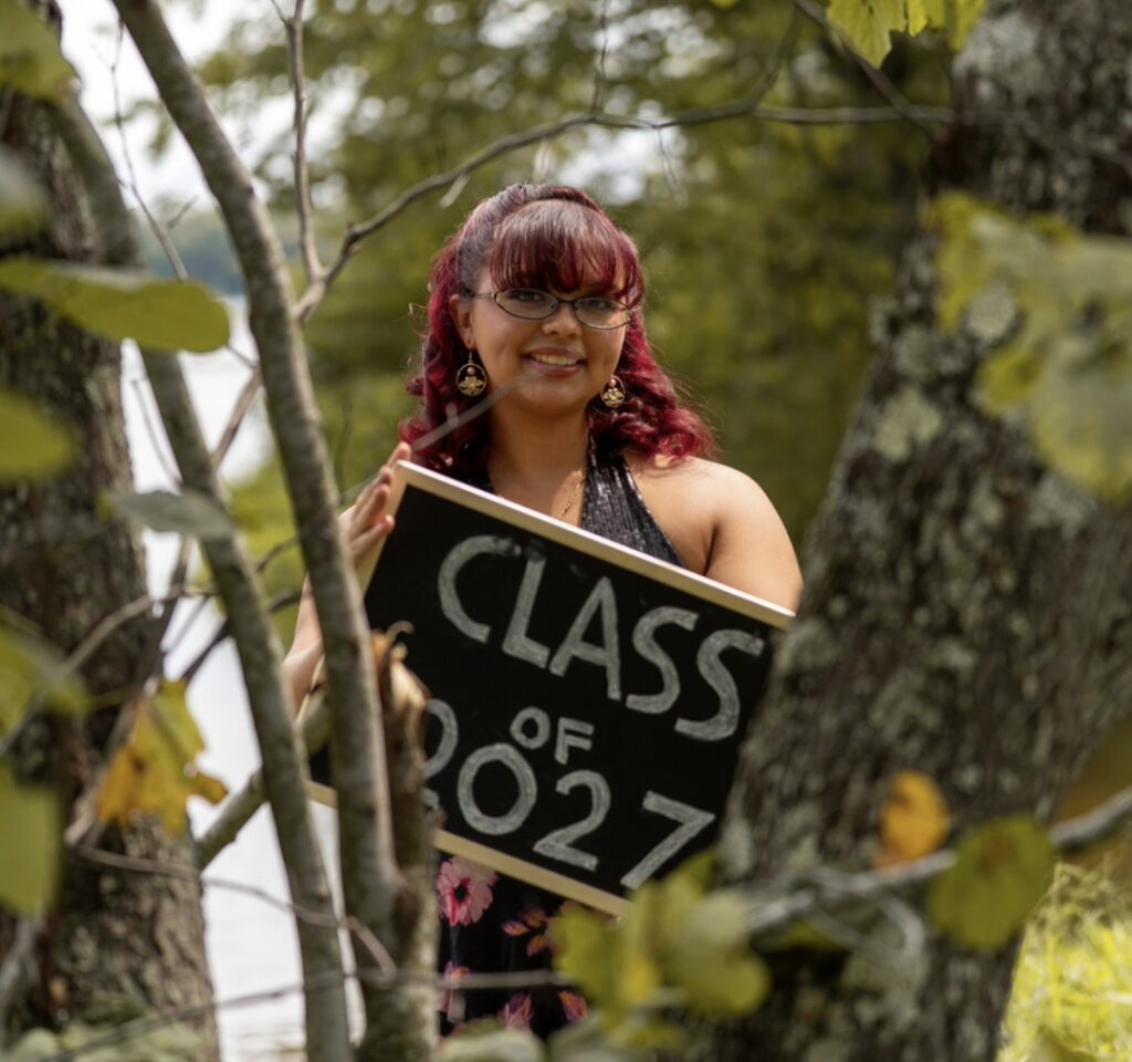 Iriellah Arbitrario our October Student of the Month posing in nature holding a class of 2027 sign.